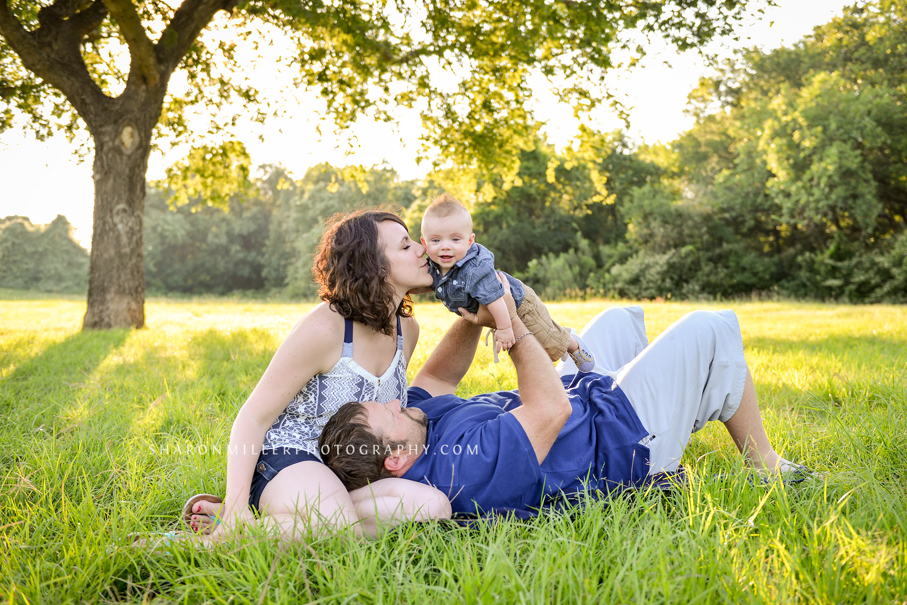 photo of Mom kissing baby being held up by Daddy under a tree by Lewisville family photographer Sharon Miller